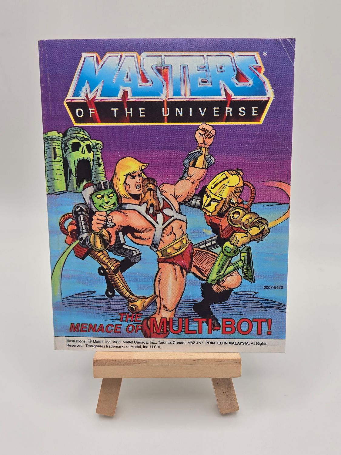 Front cover of the vintage &#39;Masters of the Universe&#39; mini comic titled &#39;The Menace of Multi-Bot!&#39; displayed on a small wooden easel. The cover features He-Man battling the robotic Multi-Bot, with Castle Grayskull in the background.