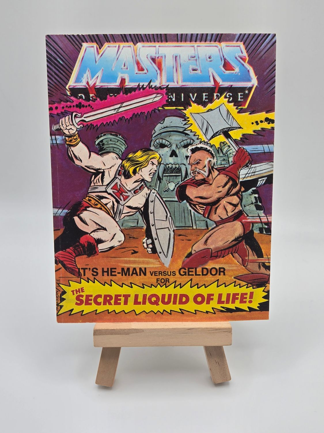 Front cover of the English version of the vintage &#39;Masters of the Universe&#39; mini comic titled &#39;He-Man versus Geldor for The Secret Liquid of Life!&#39; displayed on a small wooden easel. The cover features He-Man and Geldor in combat, with Castle Grayskull in the background.