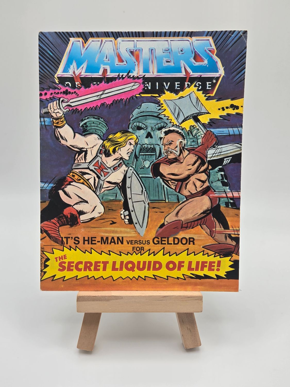 Front cover of the English version of the vintage &#39;Masters of the Universe&#39; mini comic titled &#39;He-Man versus Geldor for The Secret Liquid of Life!&#39; displayed on a small wooden easel. The cover features He-Man and Geldor in combat, with Castle Grayskull in the background.