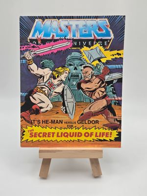 Front cover of the English version of the vintage &#39;Masters of the Universe&#39; mini comic titled &#39;He-Man versus Geldor for The Secret Liquid of Life!&#39; displayed on a small wooden easel. The cover features He-Man and Geldor in combat, with Castle Grayskull in the background.