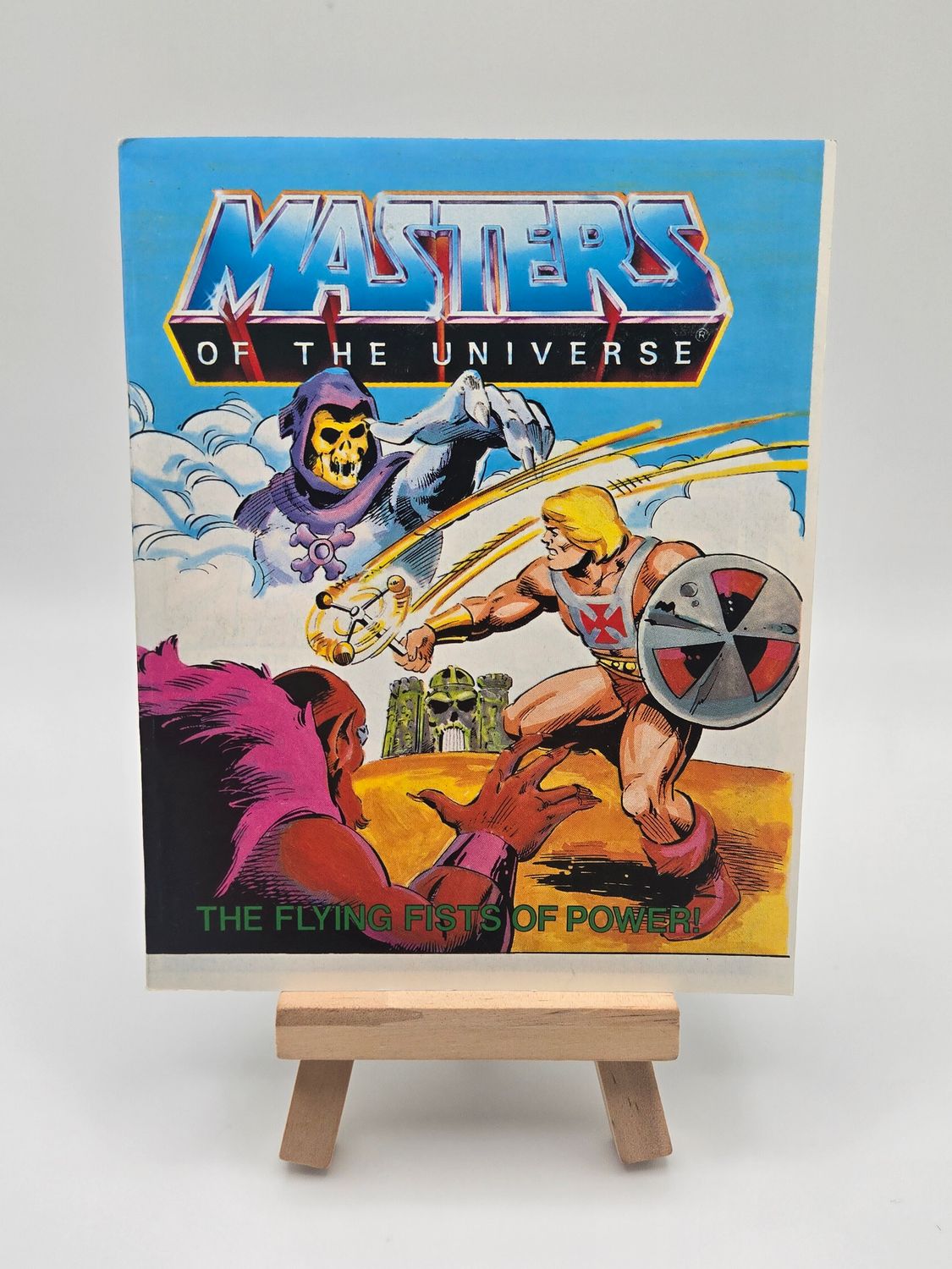 Front cover of the English version of the vintage &#39;Masters of the Universe&#39; mini comic titled &#39;The Flying Fists of Power!&#39; displayed on a small wooden easel. The cover features He-Man with his spinning shield and flying fists facing Skeletor, with Castle Grayskull in the background.