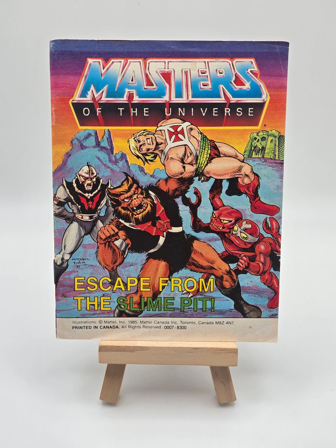 Front cover of the English version of the &#39;Masters of the Universe&#39; mini comic titled &#39;L&#39;évasion du gouffre gluant&#39; displayed on a small wooden easel. The cover shows Musclor (He-Man) captured by Hordak and other villains, with Castle Grayskull in the background.
