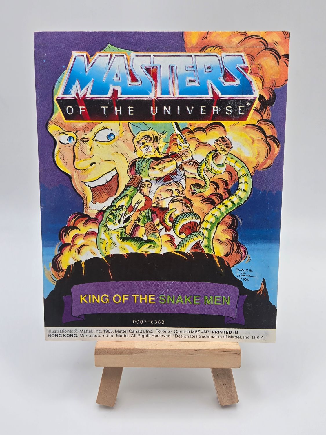 Front English cover of the 1985 &#39;King of the Snake Men&#39; - Mini Masters of the Universe (MOTU) Vintage Comic Book by Mattel, displayed on a small wooden easel, showcasing vibrant artwork of He-Man and King Hiss.