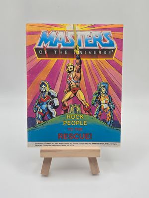 Image of the English version of vintage &quot;Les Maîtres de l&#39;Univers&quot; mini comic book titled &quot;He-Man contre Skeletor dans le Temple des Ténèbres!&quot; on a wooden display stand. The cover depicts He-Man with his sword raised, facing Skeletor, against a colorful, energy-filled backdrop.
