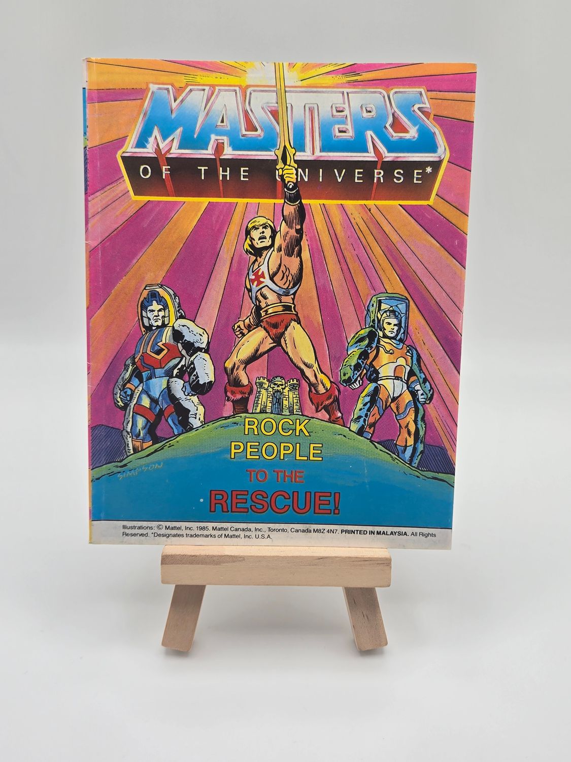Image of the English version of vintage &quot;Les Maîtres de l&#39;Univers&quot; mini comic book titled &quot;He-Man contre Skeletor dans le Temple des Ténèbres!&quot; on a wooden display stand. The cover depicts He-Man with his sword raised, facing Skeletor, against a colorful, energy-filled backdrop.