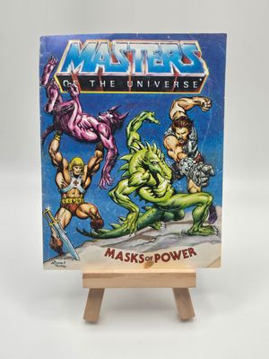 Display of the English version of the Masters of the Universe mini comic, &quot;Masks of Power&quot; on a wooden stand, depicting Musclor (He-Man) and allies in an action-packed scene against formidable foes.