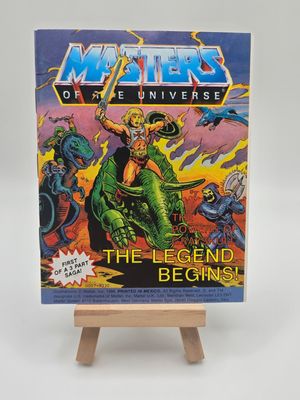 The English version of the &quot;Masters of the Universe&quot; mini comic displayed on a small wooden easel, titled &quot;The Powers of Grayskull: The Legend Begins!&quot;, showcasing vibrant cover art with He-Man wielding a sword atop a green dragon, marked as the first part of a three-part saga.