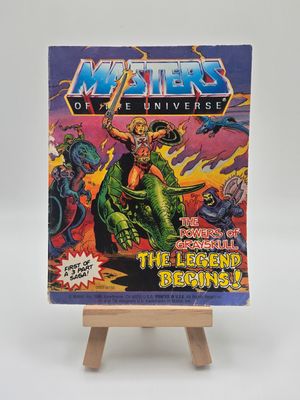 A vintage &quot;Masters of the Universe&quot; mini comic book titled &quot;The Powers of Grayskull: The Legend Begins!&quot; displayed on a small wooden easel. The cover shows He-Man with a sword atop a green beast, surrounded by enemies and dramatic skies, emphasizing the comic&#39;s epic narrative.