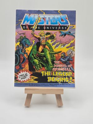 A vintage &quot;Masters of the Universe&quot; mini comic book titled &quot;The Powers of Grayskull: The Legend Begins!&quot; displayed on a small wooden easel. The cover shows He-Man with a sword atop a green beast, surrounded by enemies and dramatic skies, emphasizing the comic&#39;s epic narrative.