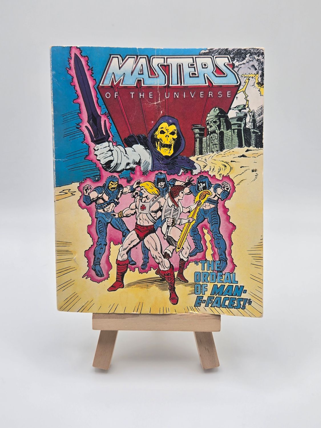 A front view of the &quot;Masters of the Universe&quot; comic book titled &quot;The Ordeal of Man-E-Faces,&quot; displayed on a wooden easel. The cover features vibrant comic artwork of He-Man and his allies battling Skeletor, set against a dramatic backdrop. 