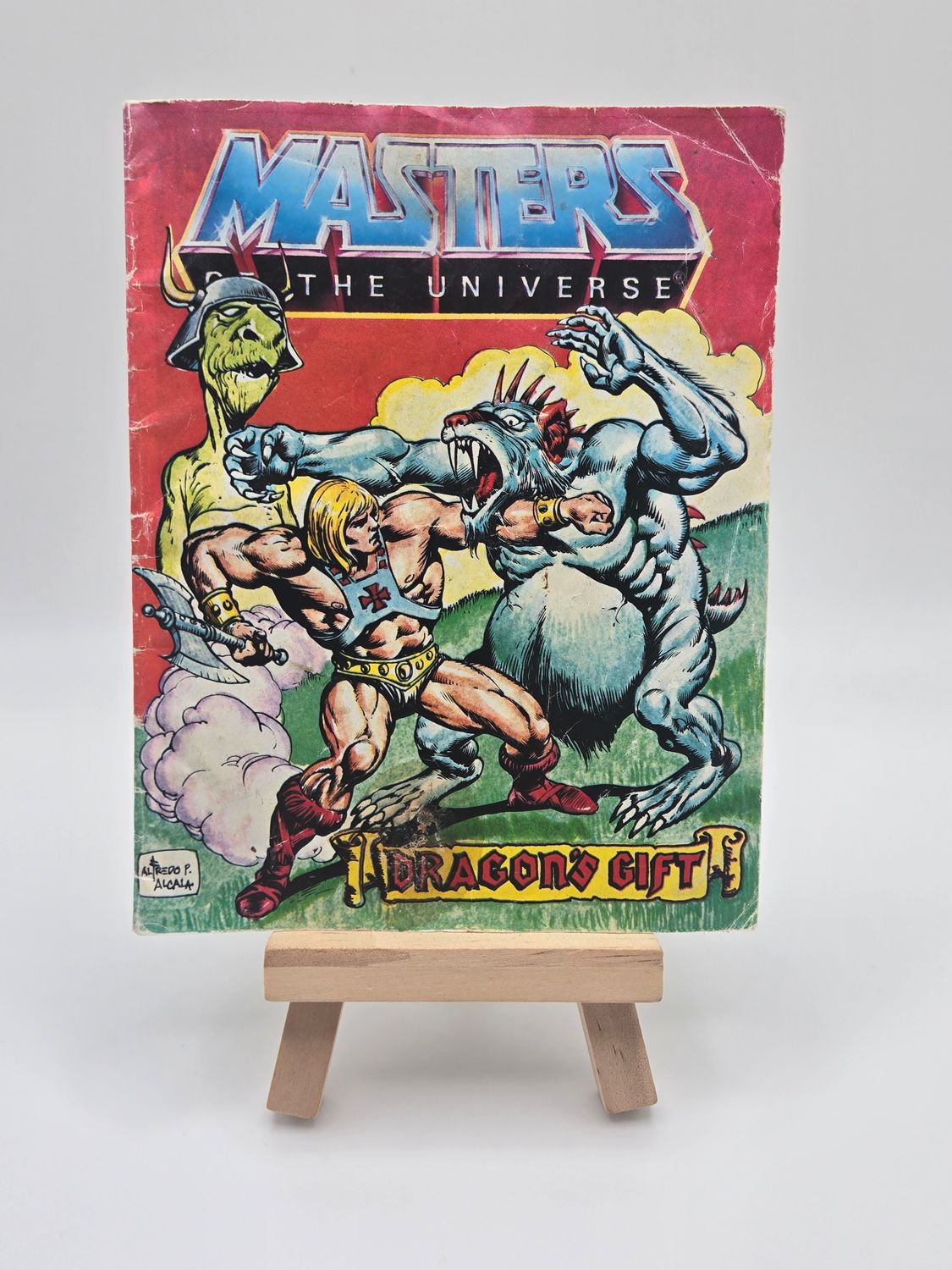 Front view of the &quot;Masters of the Universe&quot; comic book &quot;Dragon&#39;s Gift&quot; on a wooden easel. The cover features colorful artwork with He-Man and other characters engaged in intense action.