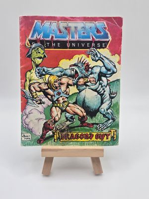 Front view of the &quot;Masters of the Universe&quot; comic book &quot;Dragon&#39;s Gift&quot; on a wooden easel. The cover features colorful artwork with He-Man and other characters engaged in intense action.