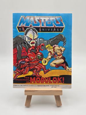 Front view of the &quot;Masters of the Universe&quot; comic book titled &quot;The Treachery of Modulok!&quot; on a wooden easel. The cover features dynamic artwork of He-Man in combat with Modulok.