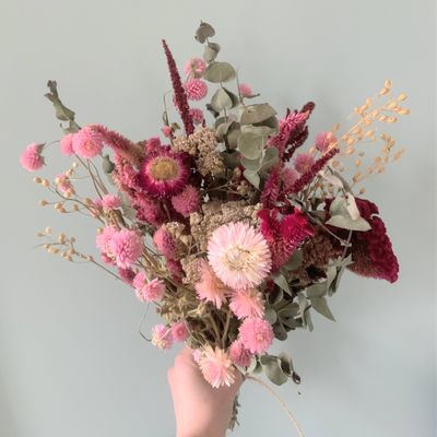 Pink and green dried flowers arranged in a bouquet, held in front of a white background.