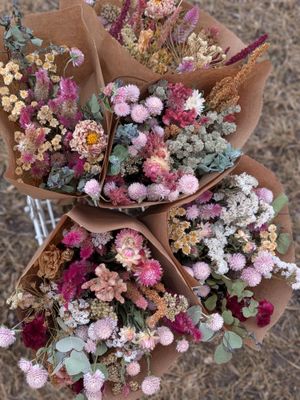 Several pink and green dried flower bouquets in brown paper wraps in a white basket in front of a tan background.