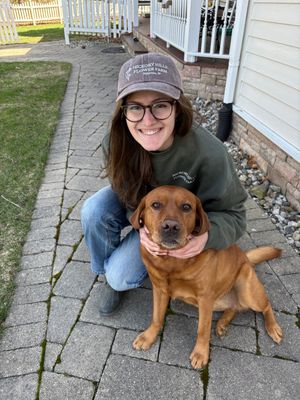 Woman wearing grown hat with white logo, standing with dog on a sidewalk.