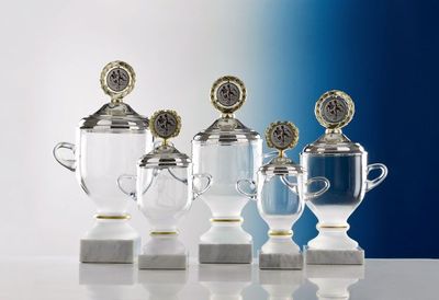 Glaspokal Glaspokal „Cremona“ mit klarem Glaskorpus, Goldrand, Deckel und Kranz auf weißem Marmorsockel.