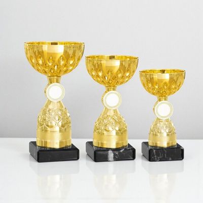 Pokal Mila mit strukturierter goldfarbener Schale, rundem Emblemträger, dekorativem Mittelteil und schwarzem Marmorsockel