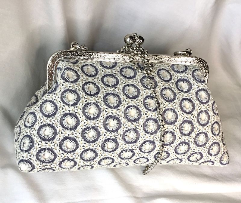 Bolso de batista francesa bordado entono gris claro y blanco la boquilla es plateada de metal y cosida a mano, el forro es amarillo con el tapacosturas azul claro con topitos blancos, tiene un bolsillo las medidas: Al15cntX30 de AnX10 de Fon la cadena es metálica de color plateada con bolitas como eslabones.
