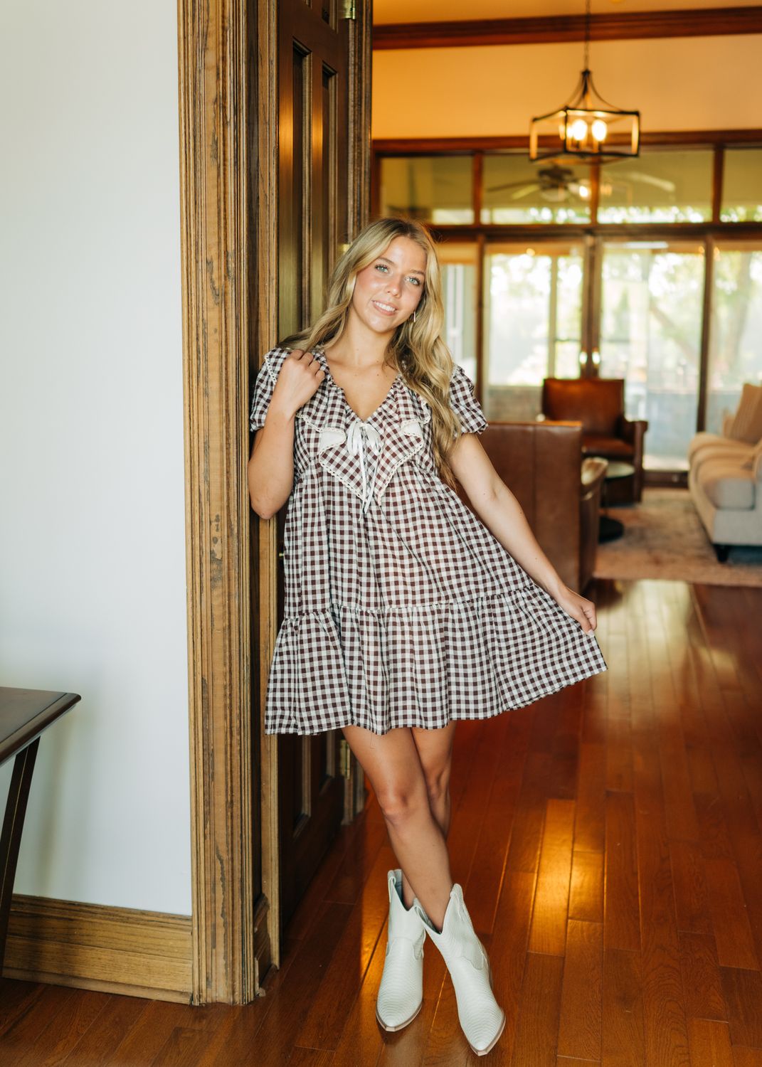 Brown gingham babydoll dress styled with white boots, model holding skirt out slightly.