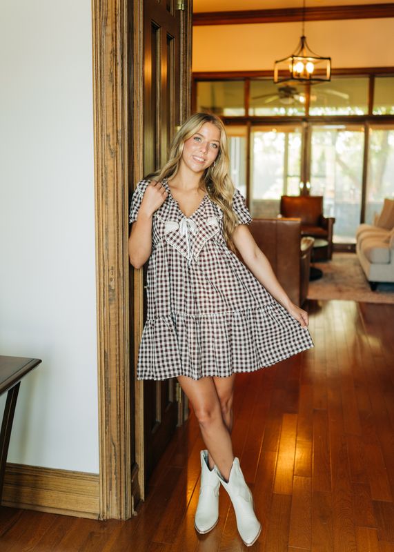 Brown gingham babydoll dress styled with white boots, model holding skirt out slightly.