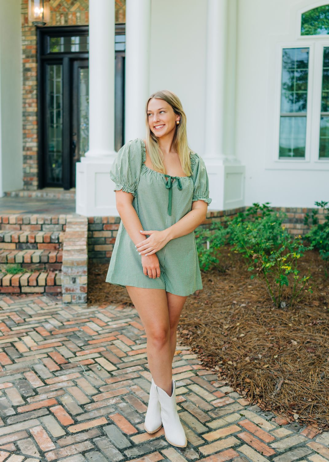 Smiling woman modeling green gingham romper with puff sleeves and tie front, hands crossed.