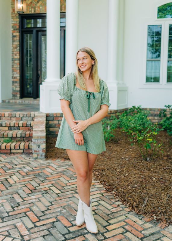 Smiling woman modeling green gingham romper with puff sleeves and tie front, hands crossed.