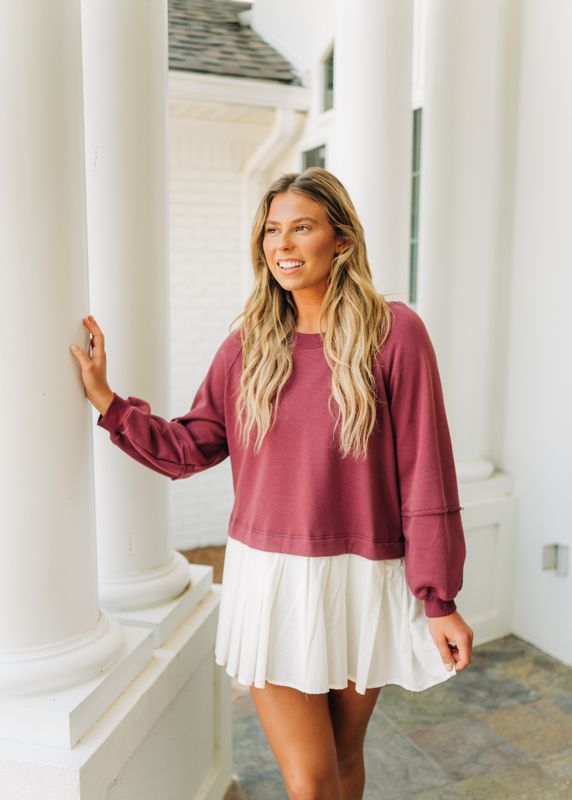 Model smiling in a plum sweatshirt dress with white pleated hem, leaning against white columns for a feminine lifestyle look.