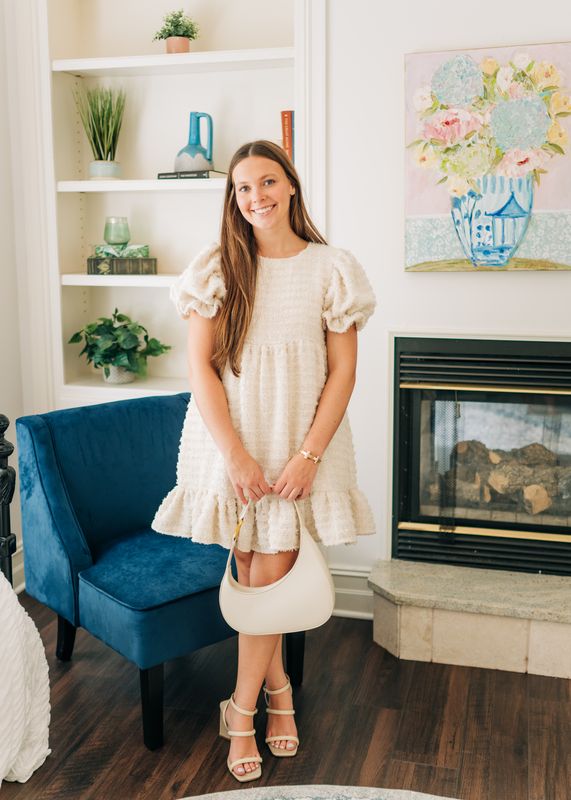 Woman wearing cream textured puff sleeve mini dress holding white handbag in a chic living room.