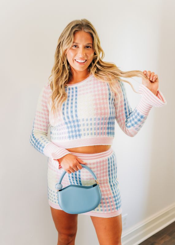 Model standing in white room wearing pastel plaid knit mini skirt in pink, blue, and yellow with white heels and blue handbag