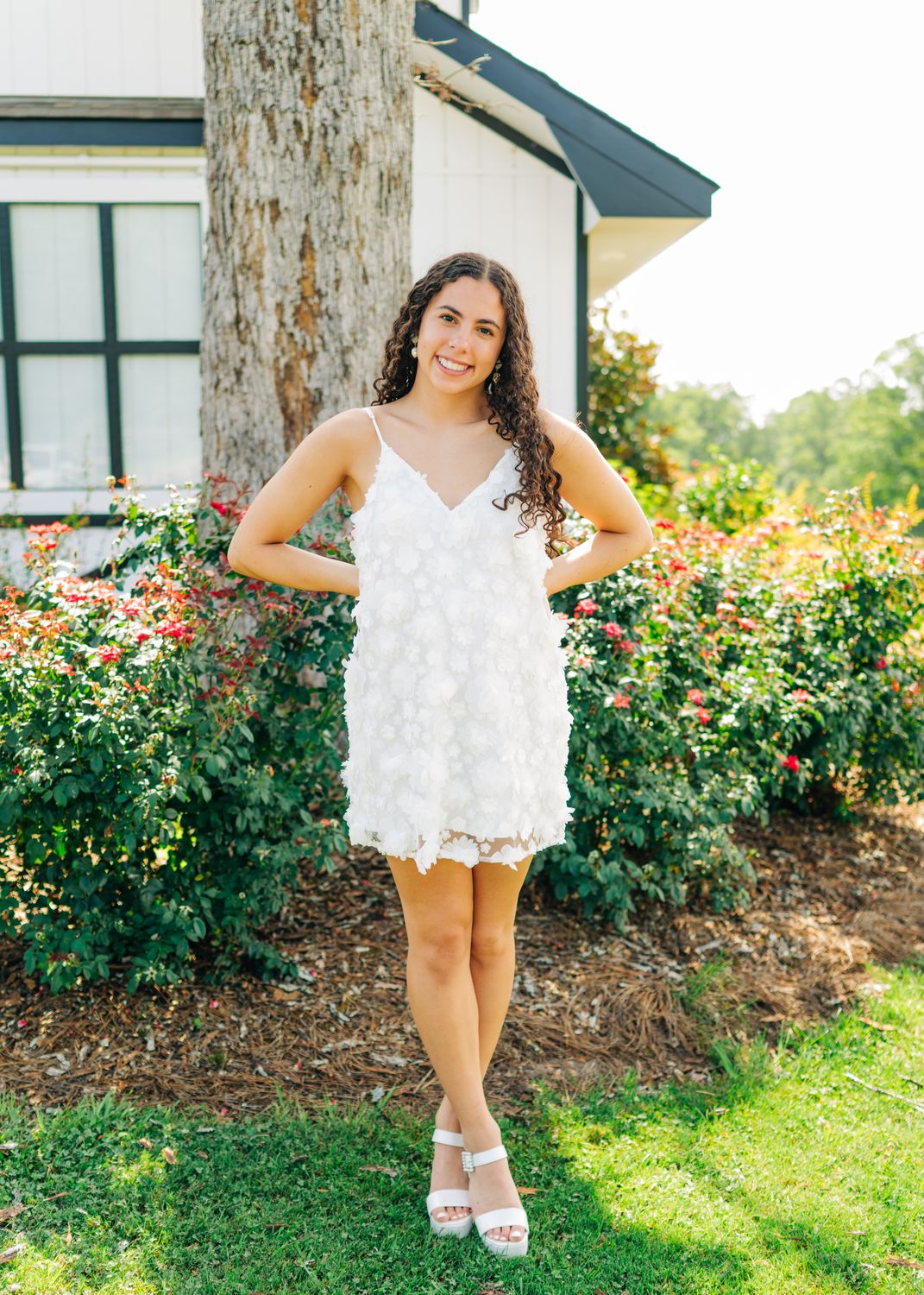 Model smiling in a white 3D floral appliqué mini dress with spaghetti straps, standing in front of greenery and flowers.
