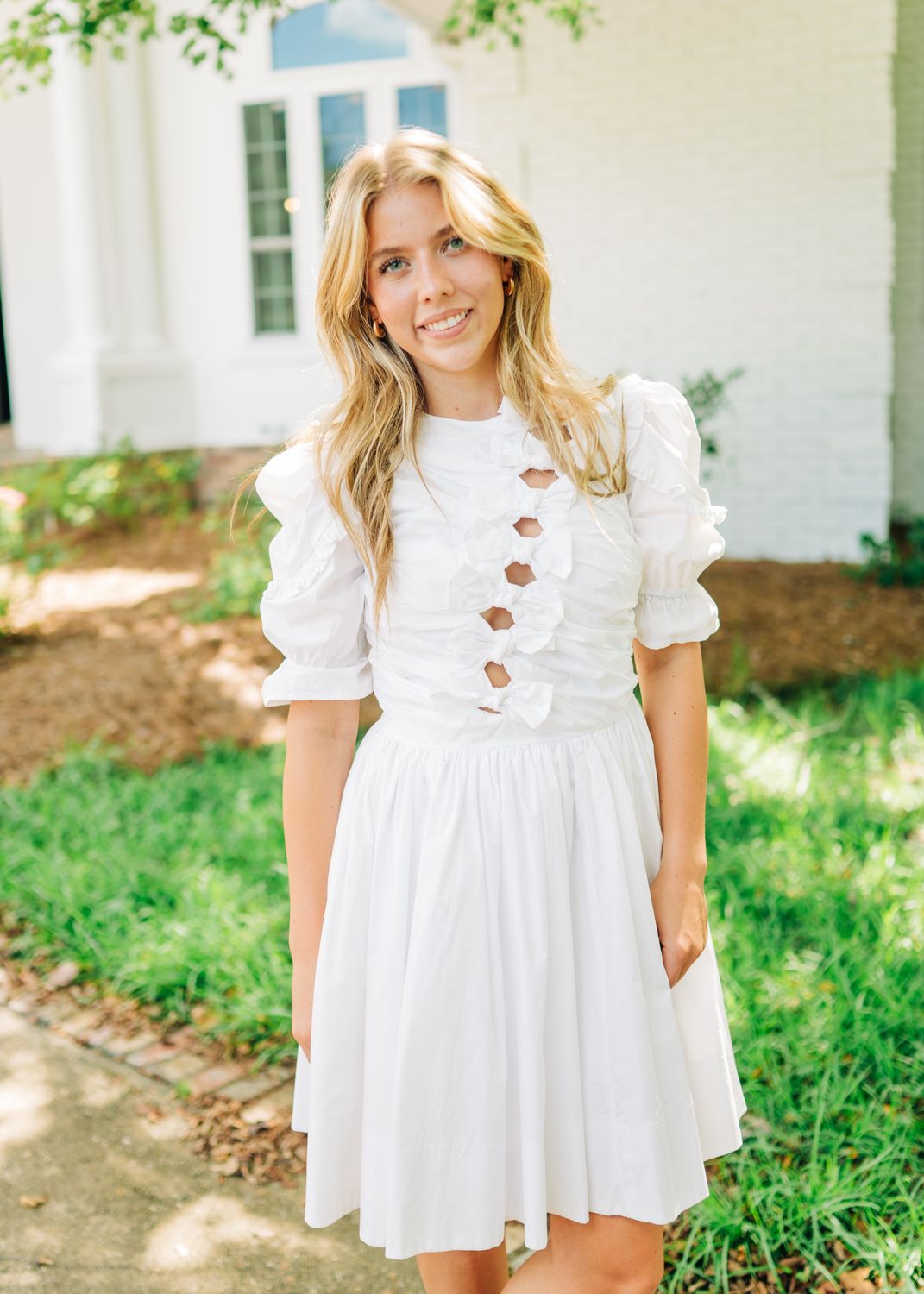 Blonde woman wearing a white puff sleeve mini dress with bow front cutouts, smiling outdoors in natural light.