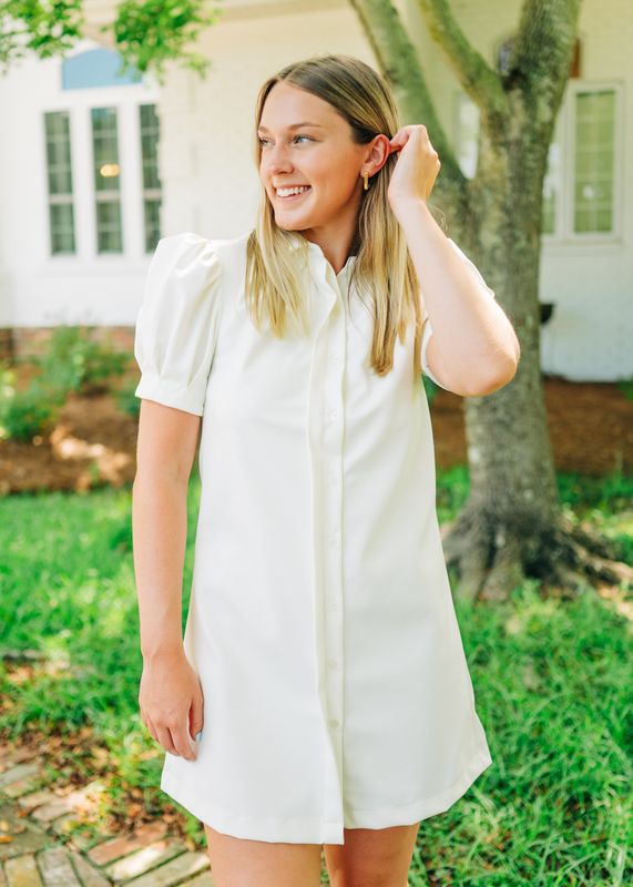  Close-up of woman wearing a white puff sleeve button-front mini dress with collared neckline, smiling outdoors by a tree.