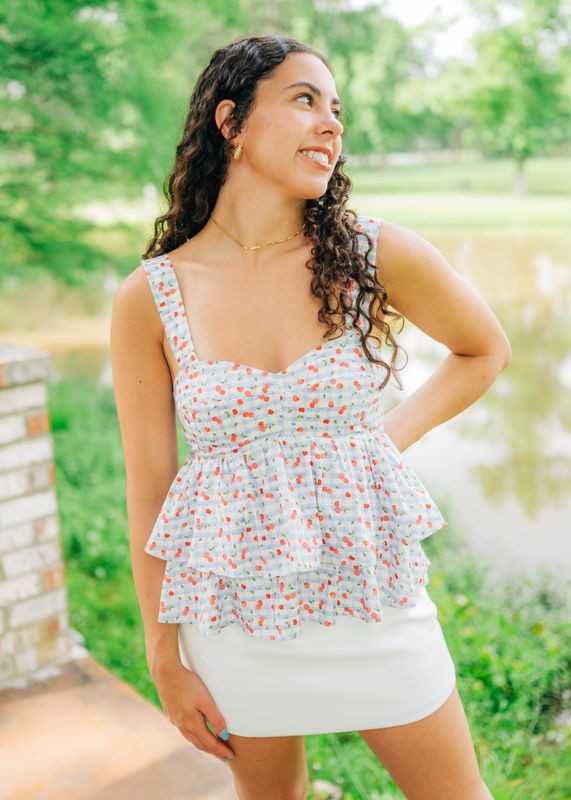White peplum tank top with cherry print, sweetheart neckline, and tiered layers, model smiling outdoors.
