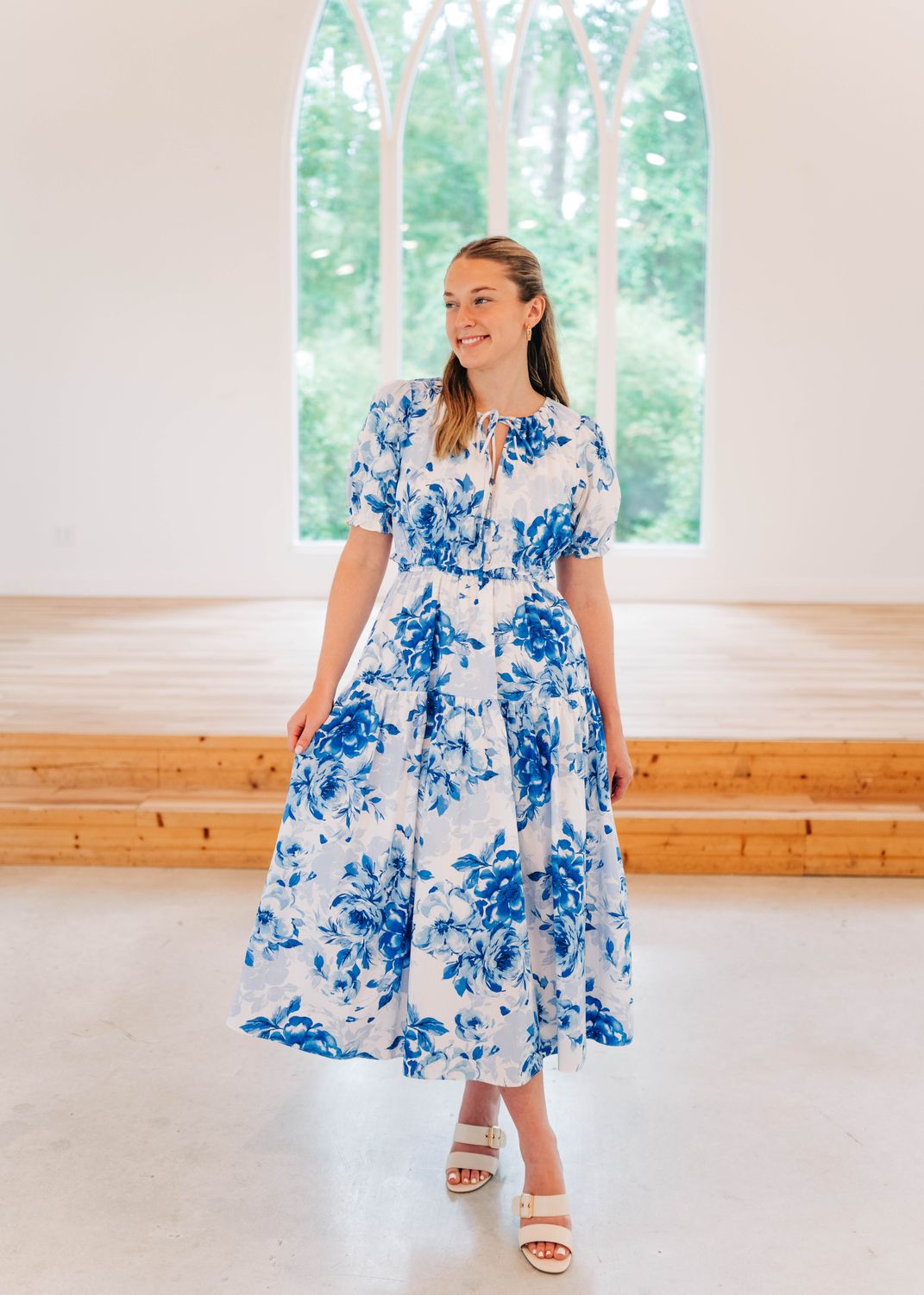 Woman wearing a blue and white floral midi dress with puff sleeves and smocked waist, styled with white heels, standing.
