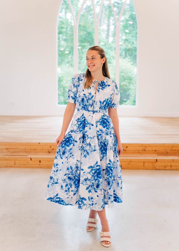 Woman wearing a blue and white floral midi dress with puff sleeves and smocked waist, styled with white heels, standing.