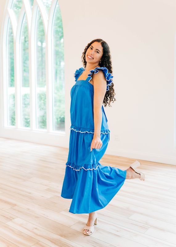 Woman twirling in royal blue ruffle sleeve tiered maxi dress with white trim, styled with nude block heels in a bright chapel