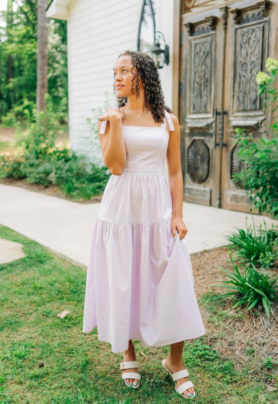 Woman in lilac tie-shoulder smocked midi dress with flowy tiered skirt, holding strap detail.