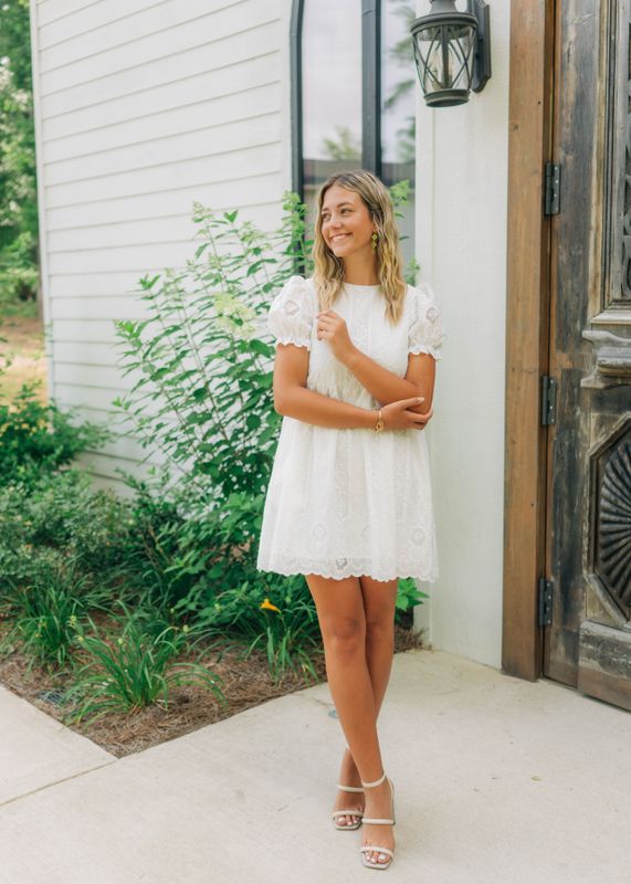 Woman wearing a white lace puff sleeve mini dress standing outside by wooden door and greenery, styled with nude heels.