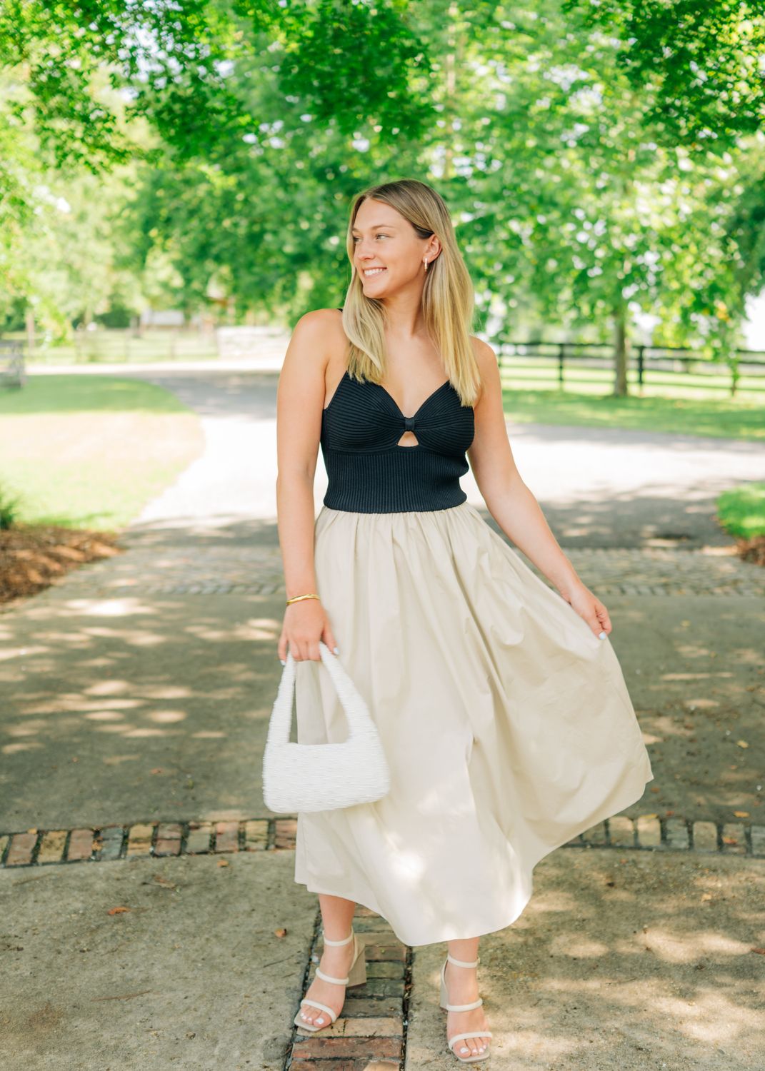 Model posing in black ribbed cutout tank dress and beige flowy midi skirt, smiling under trees.