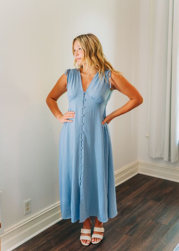 Woman wearing a light blue polka dot button-front midi dress with tie shoulder straps, styled with white block-heel sandals.