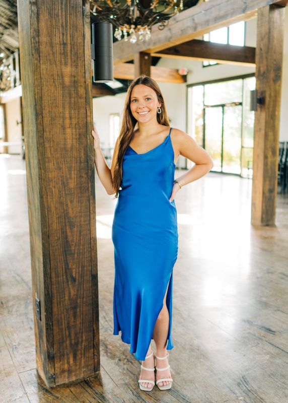 Full-body image of a woman wearing a royal blue satin slip maxi dress with cowl neckline and side slit.