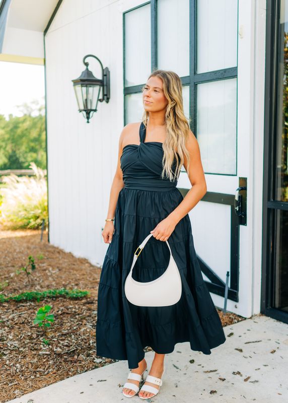 Woman wearing a black single strap tiered maxi dress, styled with white handbag and sandals, standing outdoors.