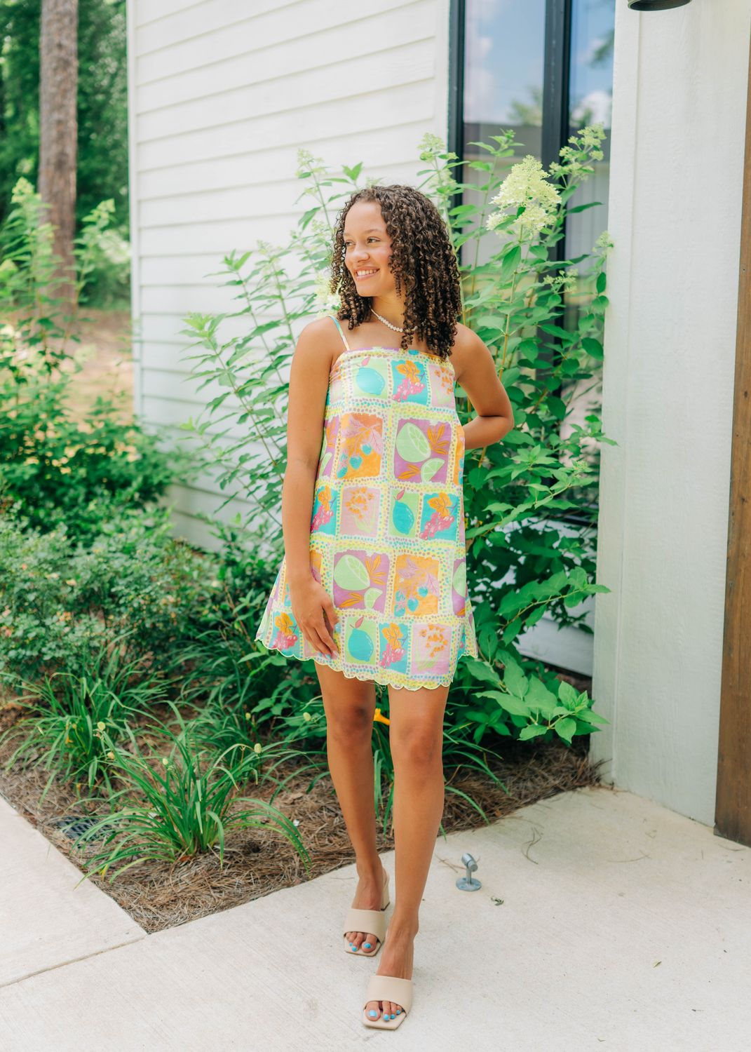 Model wearing a colorful tropical fruit print mini dress with scalloped hem and spaghetti straps, styled with nude sandals.
