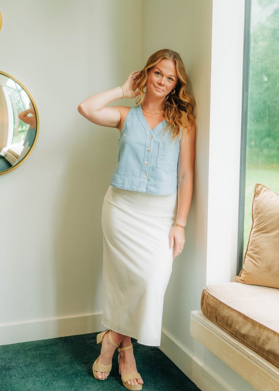 Model wearing striped denim button-front tank top with V-neckline, styled with white midi skirt and neutral block heels.