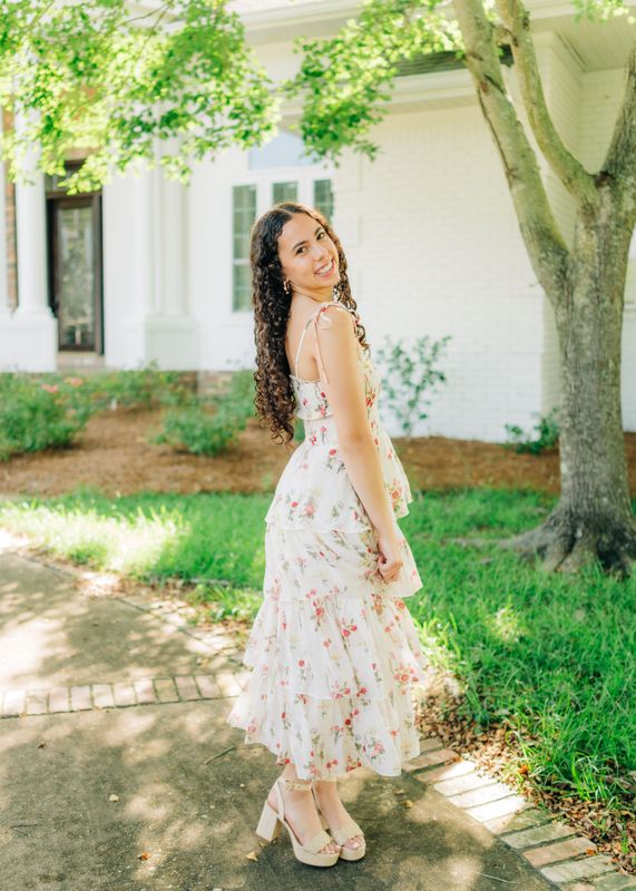 Young woman smiling over her shoulder in a white floral tiered midi dress with tie straps, standing outdoors.