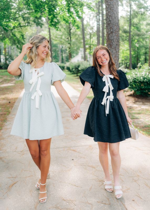 Two women holding hands wearing bow detail textured mini dresses, one in mint and one in black, styled with puff sleeves.