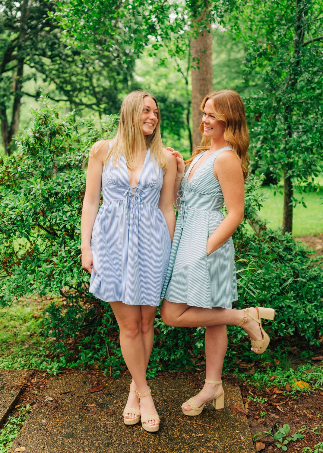 Two women wearing striped halter mini dresses in blue and green, posing outdoors in summer boutique outfits.
