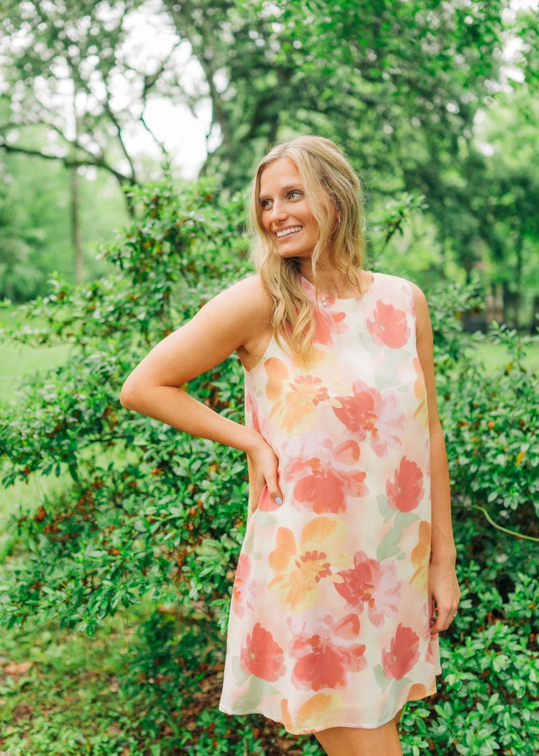 Model wearing sleeveless shift dress with watercolor floral print in coral and peach, posing outdoors.
