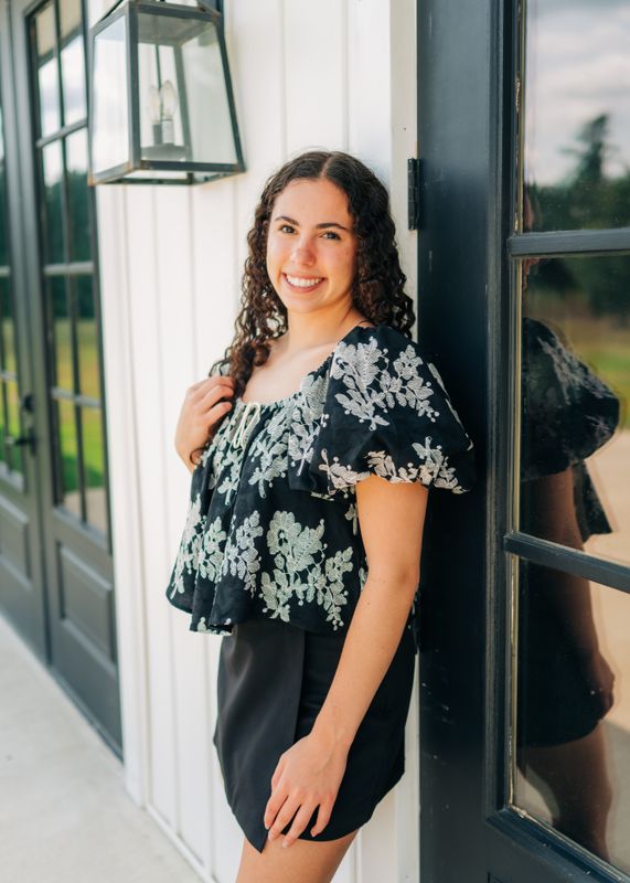 Model in black puff sleeve embroidered floral top leaning against white wall with soft smile.